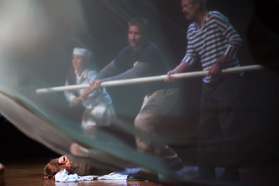 A boy in a white sailor outfit, a man in sweatshirts and cargo pants, and a man in striped blue shirt hold a white pole, someone is lying on a sheet on the ground, and a while film flutters over this whole scene in the foreground